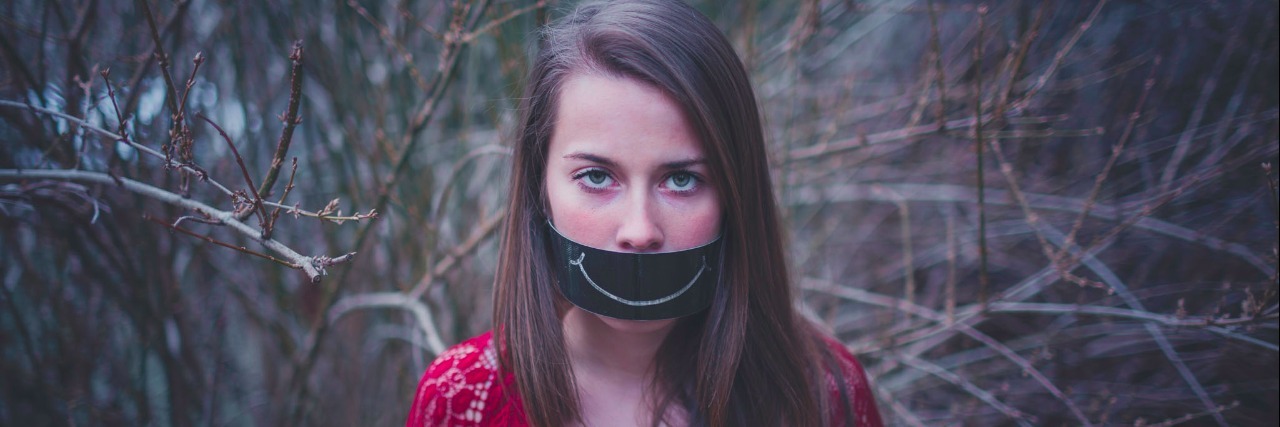 woman standing in trees with black paper over her mouth that has a white smiling mouth drawn on it