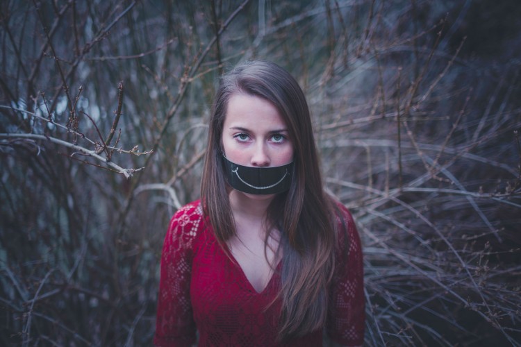 woman standing in trees with black paper over her mouth that has a white smiling mouth drawn on it