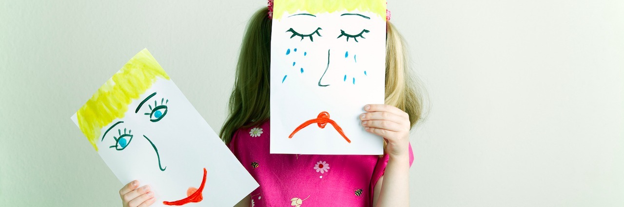 A little girl holding up a drawing of a sad face over her face