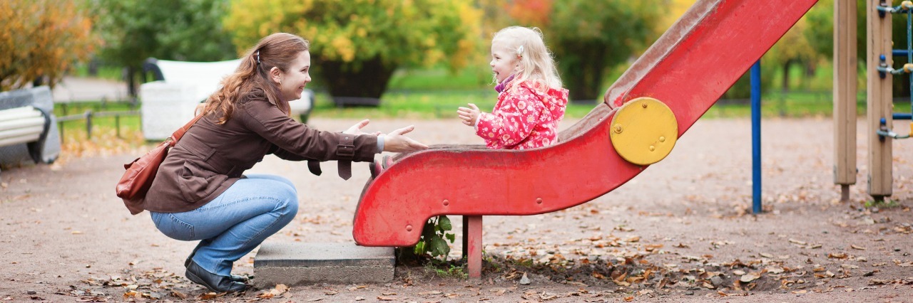 Finding Beauty in Life and Feeling Thankful for Muscle Dysfunction mother stands at the end of a slide with arms outstretched toward her daughter at the playground