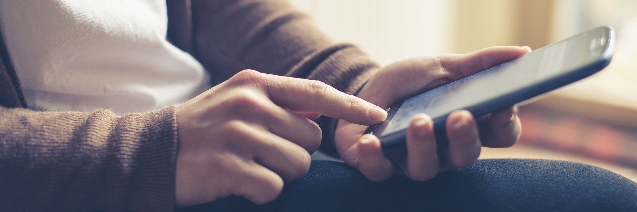 woman sitting at home wearing a brown sweater and jeans and texting on her phone