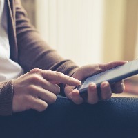 woman sitting at home wearing a brown sweater and jeans and texting on her phone