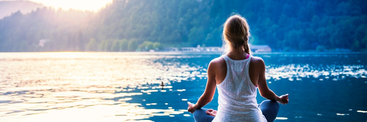 young woman meditating on a dock in front of a lake and mountains