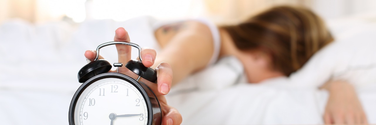 woman laying in bed with her face buried in her pillow reaching out to shut off her alarm clock