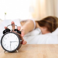 woman laying in bed with her face buried in her pillow reaching out to shut off her alarm clock