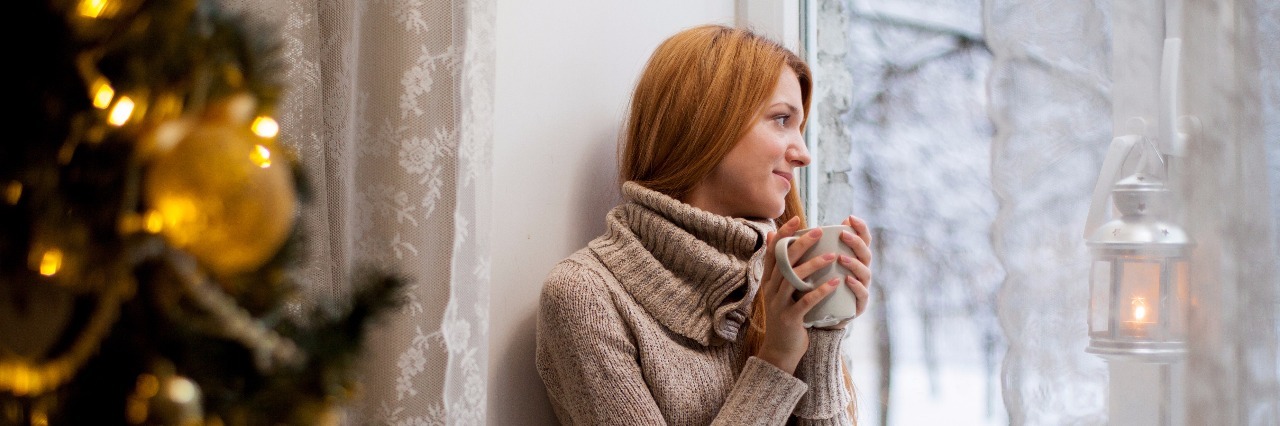 woman in brown sweater sits on a couch next to a christmas tree drinking tea and looking out a window at the snow