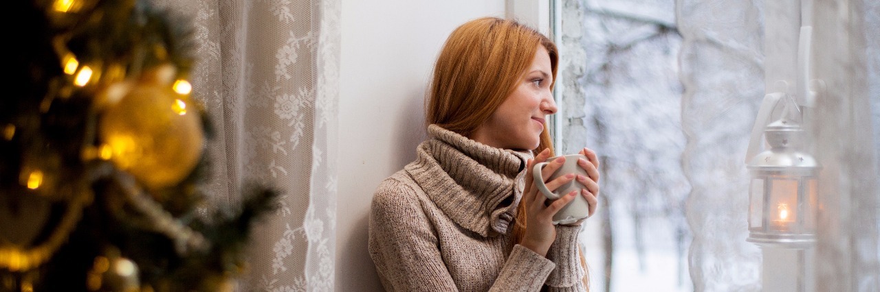 What to Consider During the Holidays With Fibromyalgia woman in brown sweater sits on a couch next to a christmas tree drinking tea and looking out a window at the snow