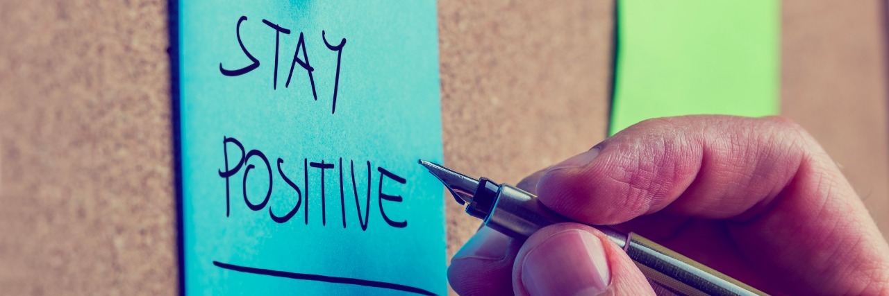 Retro image of a male hand writing Stay positive on blue post it paper pinned on cork bulletin board.