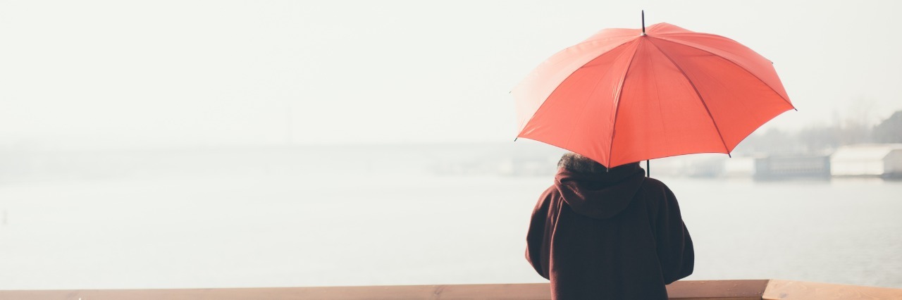 Coping With Chronic Illness and Not Feeling OK woman in dark coat standing next to railing overlooking a river and holding a red umbrella