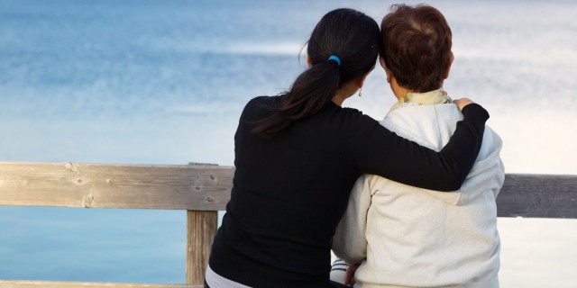 Closeup of mother with mature daughter holding her while sitting on wooden bench looking outward at the lake