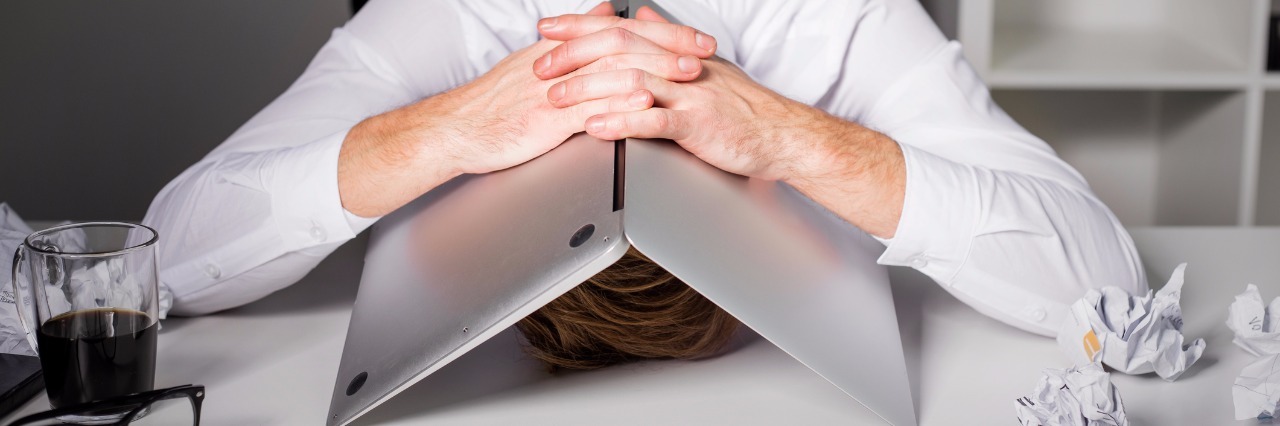 a man seated at his desk hiding his head under a laptop