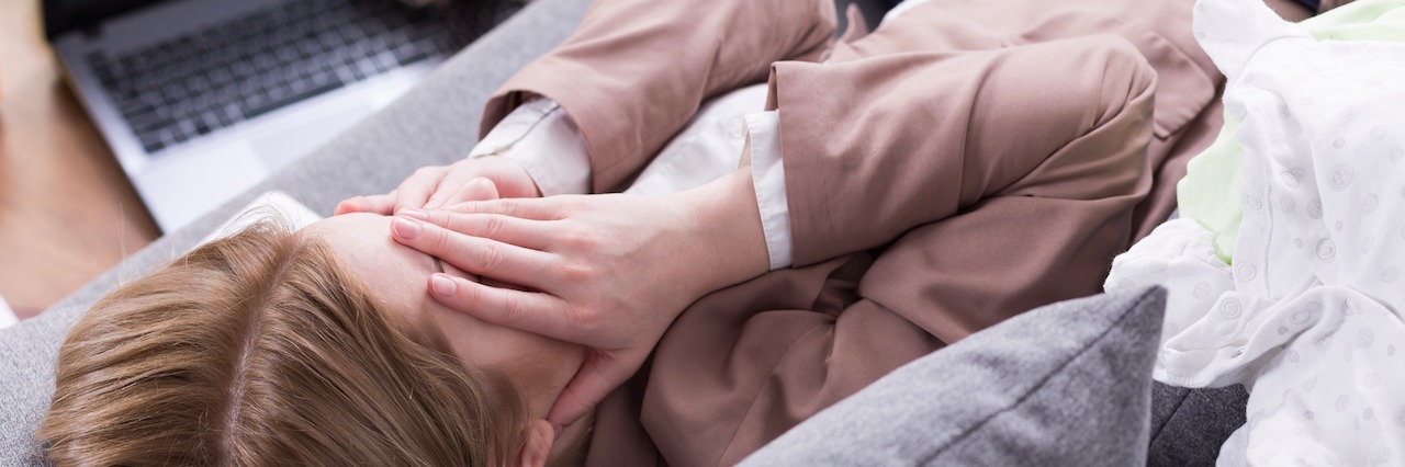 What It Is Like Parenting With Anxiety A woman laying on a couch, with her head in her hands, surrounded by toys on the floor