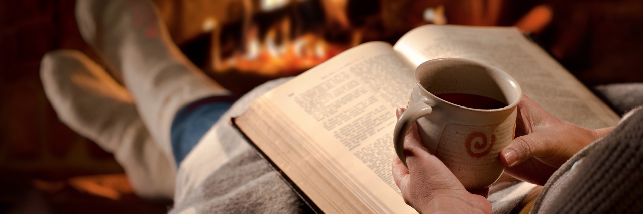 Getting Through Fatigue With Phrase 'This Too Shall Pass' person wrapped in a blanket and reading a book, holding a mug, and sitting in front of a fireplace