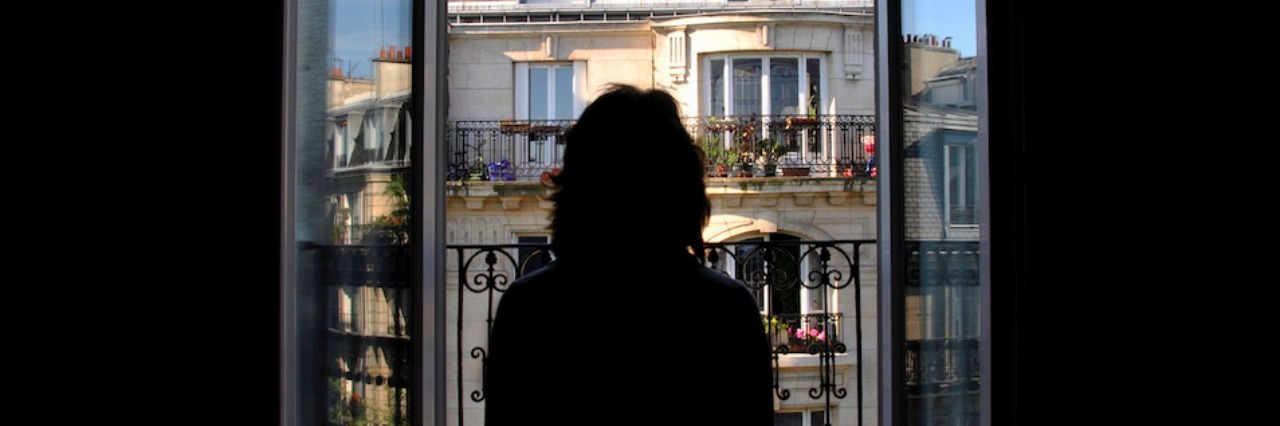 Leaving Your House for The First Time After Anxiety Kept You Indoors A woman in a dark room looking out a window