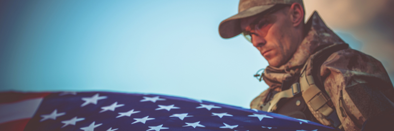 A Letter to Veterans Who Stay Silent About Their PTSD Veteran wearing camoflauge hat and clothes, holding an American flag