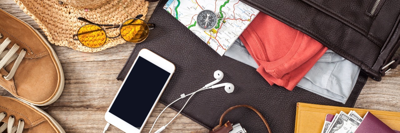 5 Tips for Traveling With Fibromyalgia and Chronic Pain suitcase filled with maps on a table with an iphone, headphones, camera, hat, sunglasses, shoes, and money