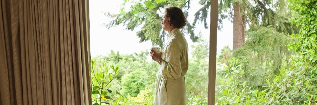 older woman wearing a bathrobe and holding a mug looking out her window at greenery