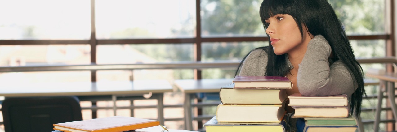 female high school student leaning on a table and gazing out a window next to a large stack of books