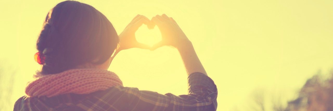 I'll Try to Be Kinder to Myself About My Chronic Illness woman making heart symbol as the sun sets