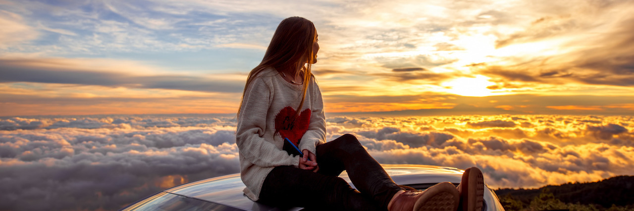 Young woman in sweater with heart shape enjoying beautiful cloudscape sitting on the car roof above the clouds on the sunrise