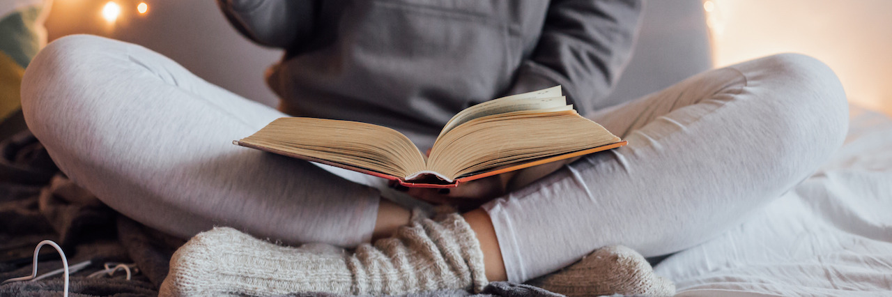 12 Tips on How to Manage Mental Illness During the Holidays Girl holding cup of hot tea and reading in bed. Around her in bad earphones, book, smart phone. Decorative lights in background.