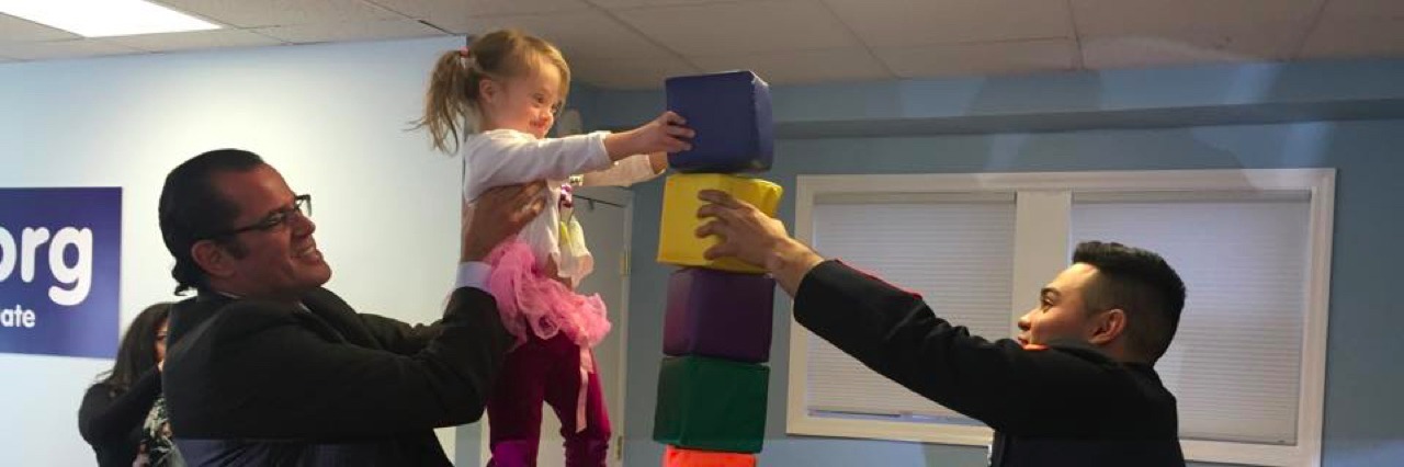 Young girl with a disability stacking blocks with a marine, her dad is lifting her to the top of the stack.