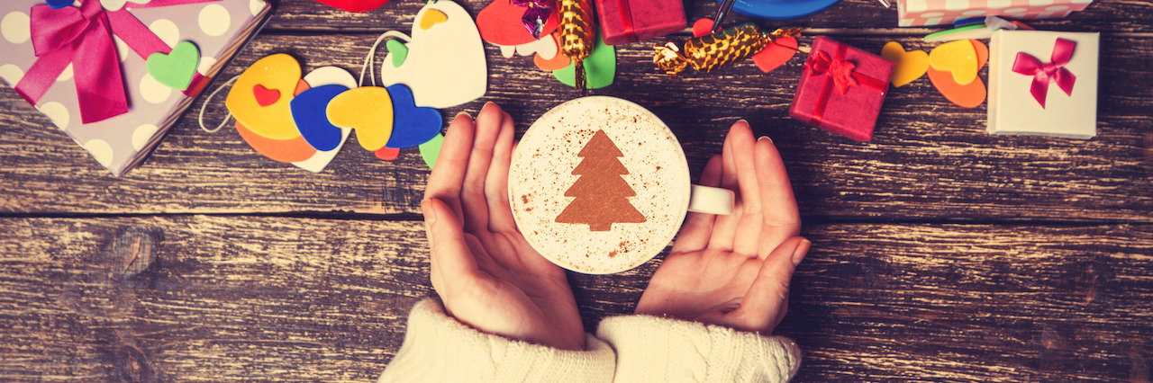 Female holding cup of coffee with cream christmas tree on a table near toys.