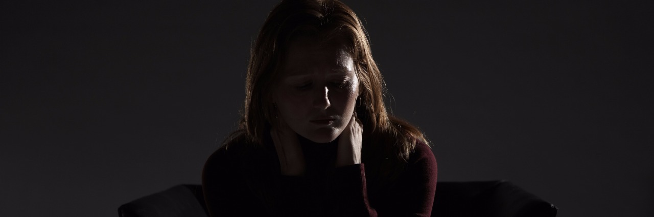 The Lies Depression Makes You Believe When You're Suicidal woman sitting on armchair in the dark rubbing neck and shoulders