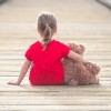 little girl in a red dress waiting on a boardwalk hugging teddybear