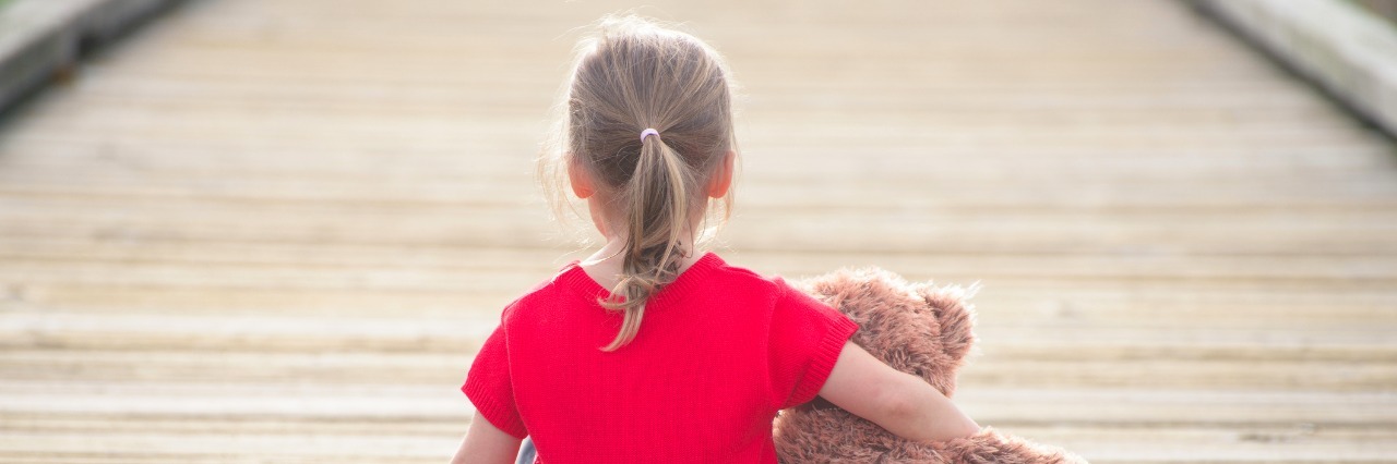 little girl in a red dress waiting on a boardwalk hugging teddybear