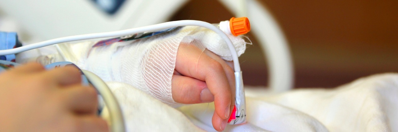 A child is laying on a hospital bed with his arm and hand raised above his head with an I.V. and bandage on it.