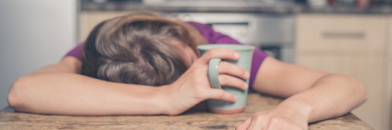 Being Tired With Myalgic Encephalomyelitis/Chronic Fatigue Syndrome a woman in a purple shirt lays her head on the table while holding a mug of tea
