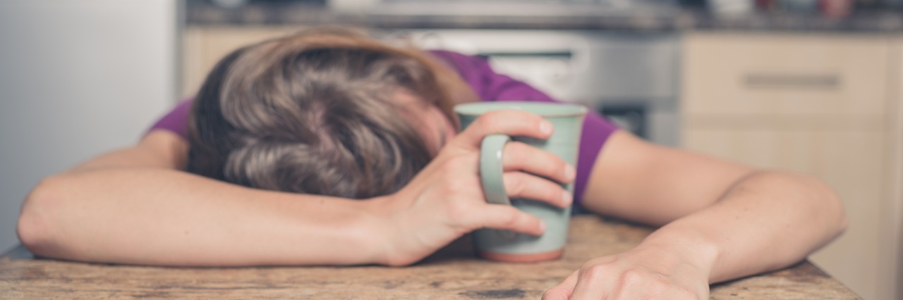 a woman in a purple shirt lays her head on the table while holding a mug of tea