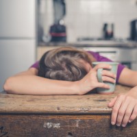 a woman in a purple shirt lays her head on the table while holding a mug of tea