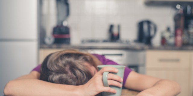 a woman in a purple shirt lays her head on the table while holding a mug of tea