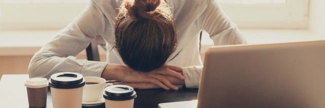 woman with her head down on her desk at work next to her laptop and several cups of coffee