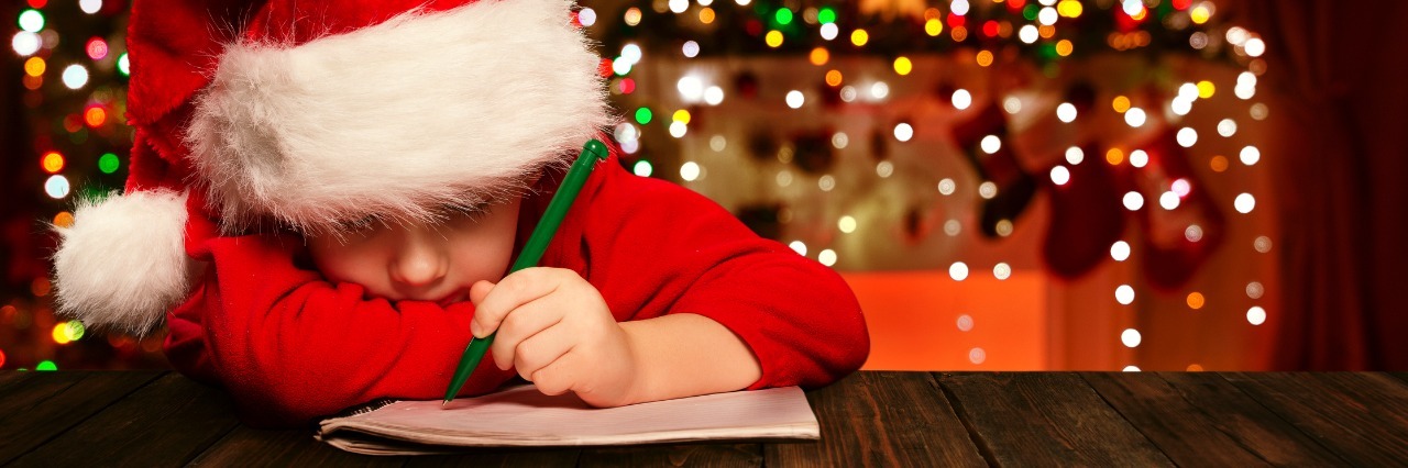 boy in santa hat writes a letter to santa with christmas decorations in the background