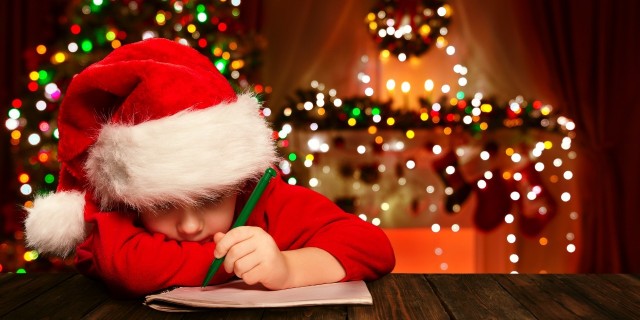 boy in santa hat writes a letter to santa with christmas decorations in the background