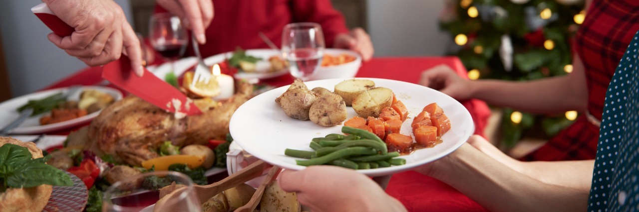Holiday Parties and Dinners With Celiac Disease woman holding out her plate of food while a family member carves the turkey. a christmas tree is in the background