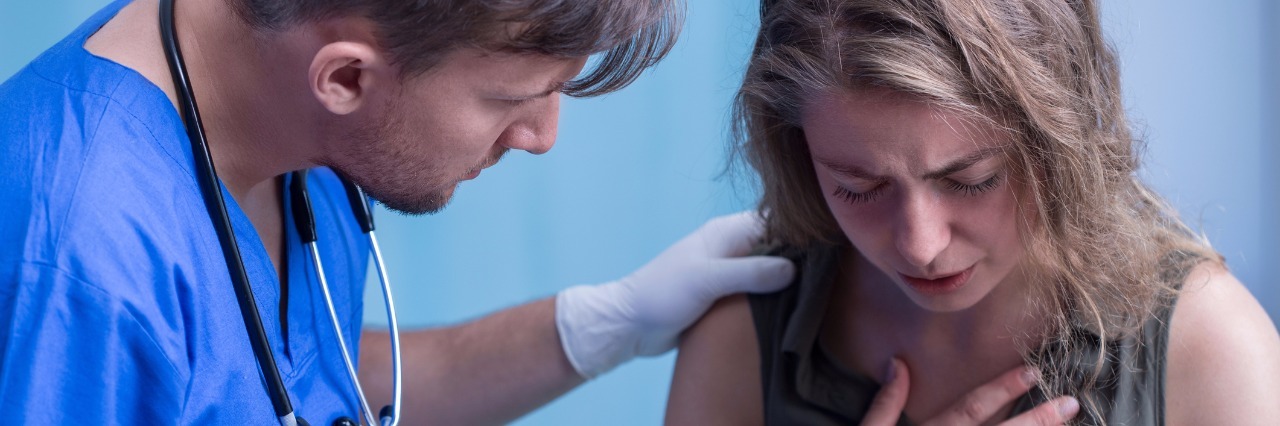What I Need From Doctors as Someone With a Rare Congenital Heart Defect young woman holds her hand over her chest in pain while a male emergency room doctor watches in concern