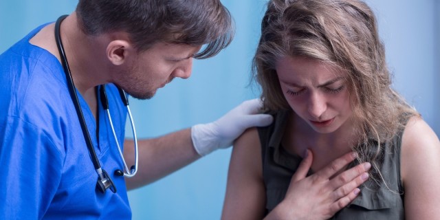 What I Need From Doctors as Someone With a Rare Congenital Heart Defect young woman holds her hand over her chest in pain while a male emergency room doctor watches in concern