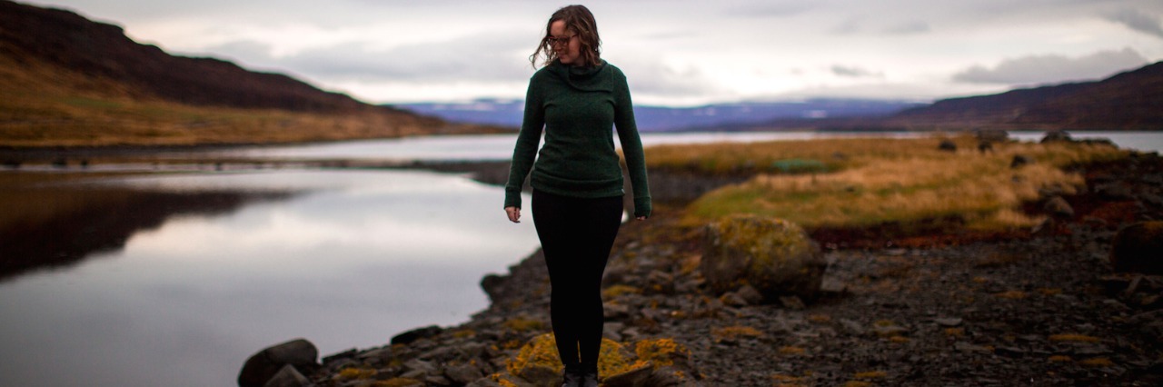 Responding to People Who Think I'm 'Weak' Because of My Mental Illness Woman, wearing a green sweater, blue hat and glasses poses on grassy land near water on an overcast day