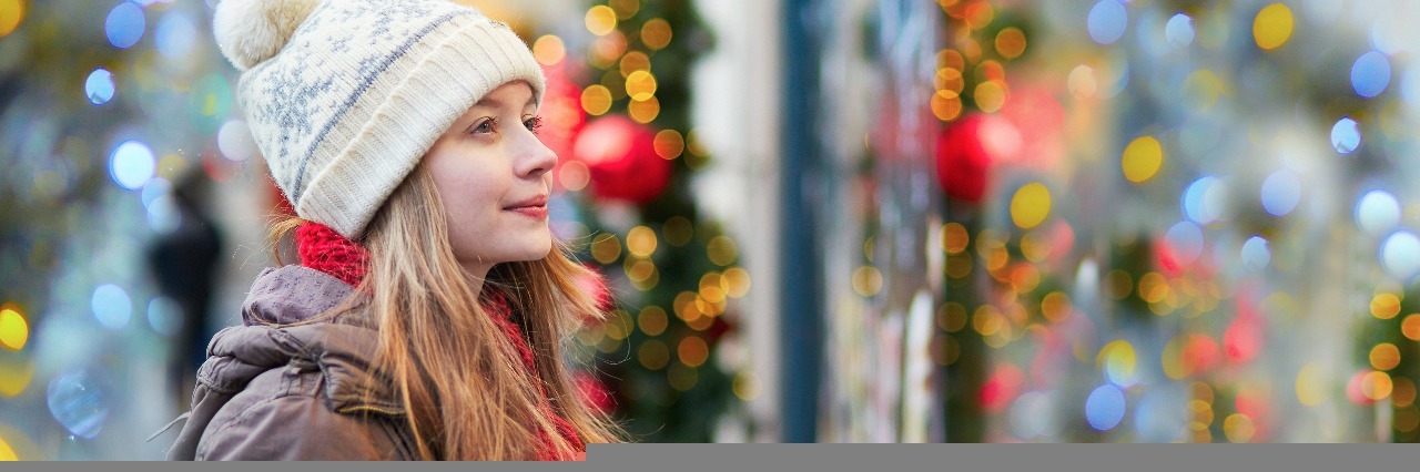 girl in a jacket and hat standing outside a store window at a christmas market