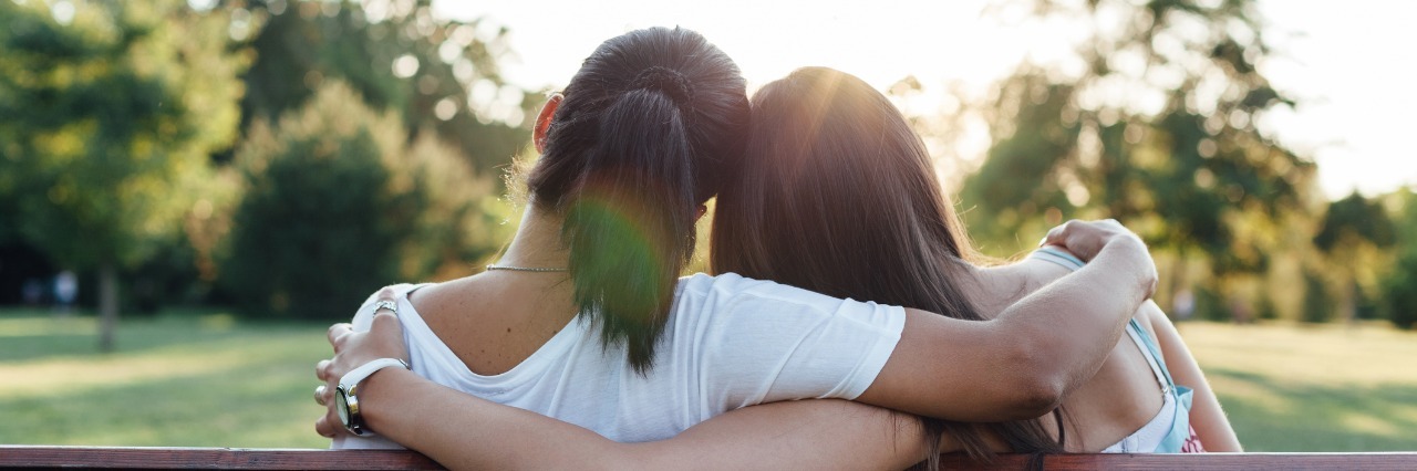 What to Know About Chronically Ill People back view of mother and daughter hugging and sitting next to each other on a park bench