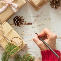 Person writing a letter to Santa on a wood table with wrapped presents on it