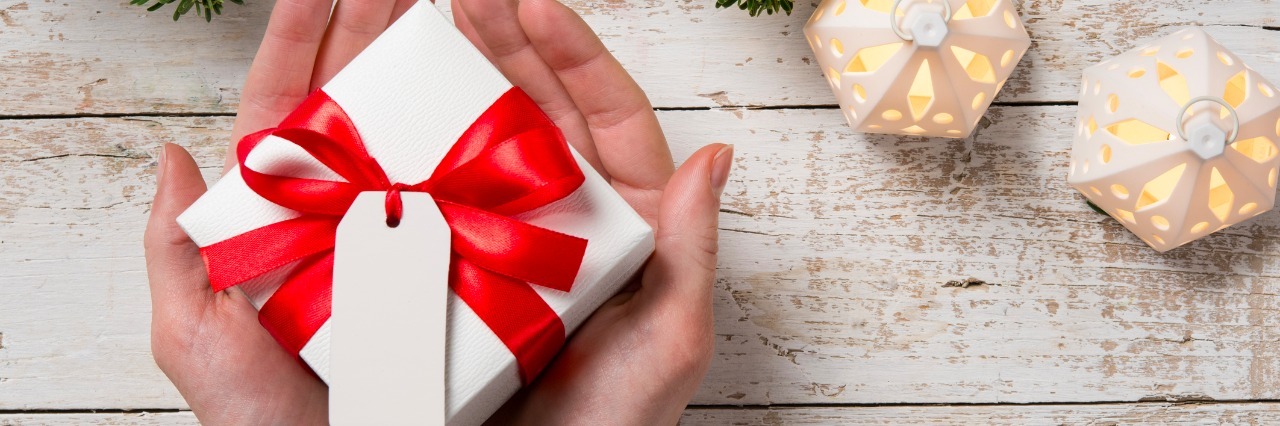 6 Great Gifts for Someone With Marfan Syndrome Top view of female hand holding white christmas gift box wrapped with red ribbon. High angle view of woman showing xmas present on rustic wooden table with fir branch on top.