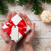 Top view of female hand holding white christmas gift box wrapped with red ribbon. High angle view of woman showing xmas present on rustic wooden table with fir branch on top.