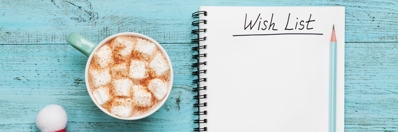 mug of hot cocoa and marshmallows, santa cap, and notebook with wish list on a blue table