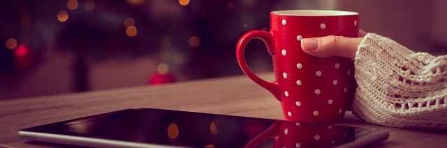 Close up of female hand holding a cup of hot coffee with tablet computer set next to it and christmas tree in the background