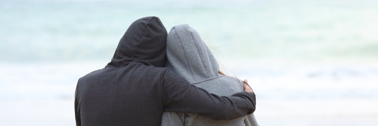 Two people wearing hoodies, hugging while facing the ocean on the beach